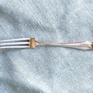 May include: A silver-toned metal fork featuring three slender tines and a smooth, tapered handle. The handle is engraved with the name Nellie in a cursive script, adding a personalized detail to the polished surface of this flatware piece.