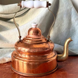 May include: A vintage copper tea kettle with a white porcelain handle and lid. The kettle has a rounded body and a spout on the side.