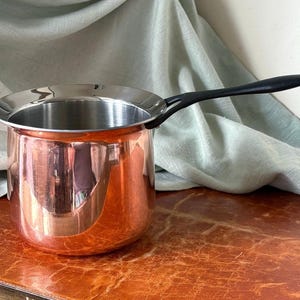 May include: A copper saucepan with a polished finish and a wide, flared silver-toned rim. The pot features a long, slender black handle extending from the side. The interior is metallic silver, and the exterior displays a warm, reflective copper hue.