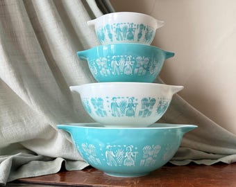 Op de afbeelding: Een set van vier in elkaar passende mengkommen met handvatten, afwisselend van wit en turkoois glas. Elke kom heeft een herhalende blauwgroene grafische print van figuren, ananassen en korenaren op de buitenkant.