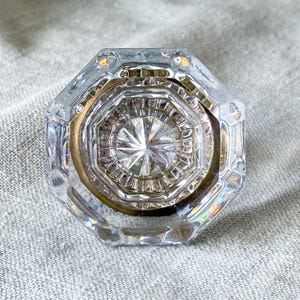 May include: A clear, faceted glass cabinet knob with an octagonal shape. The center features a starburst pattern radiating from the middle, surrounded by concentric rings and a metallic gold-toned base visible through the transparent glass.