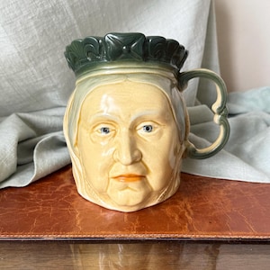 May include: A vintage ceramic mug featuring a sculpted face with a crown. The mug has a yellow face with blue eyes and an orange mouth. The handle and crown are green. The mug is likely a collectible item.