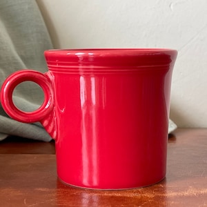 May include: A red ceramic mug with a ring handle. The mug is on a wooden surface.