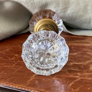 May include: A clear, multi-faceted glass door knob with a circular, scalloped edge design. The knob features a central brass-toned metal base that connects to the glass portion, showcasing a reflective, transparent surface with intricate geometric patterns.