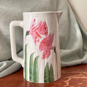 May include: White ceramic pitcher with a handle, featuring a floral design of pink flowers and green leaves.