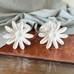 May include: Two white daisy-shaped napkin rings. Each ring features a detailed flower design with multiple petals and a textured center. The napkin rings are placed on a brown leather surface, with a light green fabric in the background.
