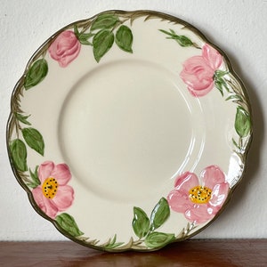 May include: A white ceramic plate with a scalloped edge features a pink rose pattern with green leaves. The plate has a cream-colored background and a gold rim.