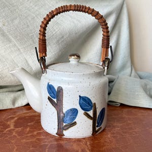 May include: A speckled white ceramic teapot with a brown woven handle and lid. The teapot features a design of brown tree branches and blue leaves. The spout and handle attachments are metal.