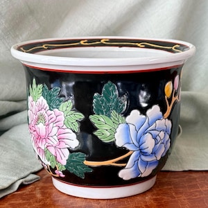 May include: Black ceramic planter with a white rim and a red accent. The planter features a floral design with pink and blue flowers and green leaves. The rim has a gold and black pattern.