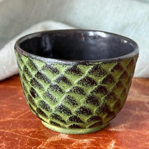 May include: A small green and black ceramic bowl with a textured, geometric pattern. The bowl is sitting on a brown surface.
