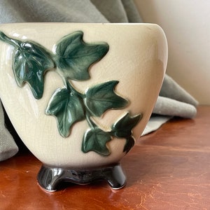 May include: A cream-colored ceramic planter with a glossy crackle glaze finish, featuring a raised, dark green ivy leaf design on the front. The vessel is supported by three short, dark brown, curved feet that elevate the base from the surface.