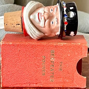 May include: A vintage porcelain bottle stopper featuring a Beefeater in a black and red hat. The stopper is sitting on a red box with the words "The Beefeater Pourer" printed in gold.