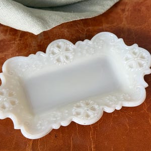 May include: A rectangular, off-white, vintage-style dish with a scalloped edge. The dish features embossed floral and geometric patterns. The background is a brown surface with a green cloth in the upper left corner.