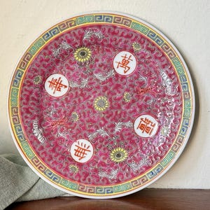 May include: A round, decorative plate with a vibrant pink background and a detailed border. The plate features floral and scroll patterns, with Chinese characters in white circles. Yellow and green accents add to the design.