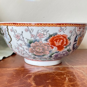 May include: A large white ceramic bowl with a floral design. The bowl features a red, orange, and pink floral pattern with green leaves and white accents. The bowl has a gold rim and a scalloped edge.