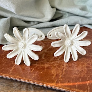 May include: Two white daisy-shaped cabinet knobs with matching backplates. The knobs have a textured, aged appearance, with a central disc and radiating petals. The knobs are displayed on a brown leather surface, suggesting a vintage or farmhouse aesthetic.