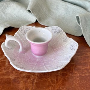 May include: A white ceramic candle holder with a pink cup and a handle. The holder has a seashell design and sits on a brown surface. A light green cloth is draped in the background.