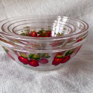 May include: A set of four clear glass nesting bowls. Each bowl is decorated with a pattern of red cherries and green leaves. The bowls are stacked inside each other, showcasing their varying sizes.