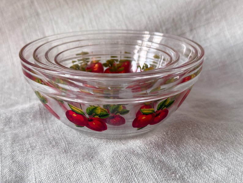 May include: A set of four clear glass nesting bowls. Each bowl is decorated with a pattern of red cherries and green leaves. The bowls are stacked inside each other, showcasing their varying sizes.
