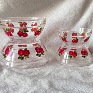 May include: Two clear glass bowls with a red apple design. The bowls are stacked, with the smaller bowl on top of the larger one. The apple design features red apples with green leaves. The bowls are on a light-colored fabric.