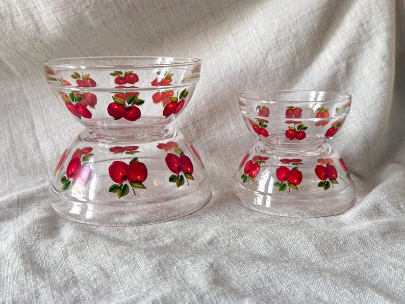 May include: Two clear glass bowls with a red apple design. The bowls are stacked, with the smaller bowl on top of the larger one. The apple design features red apples with green leaves. The bowls are on a light-colored fabric.