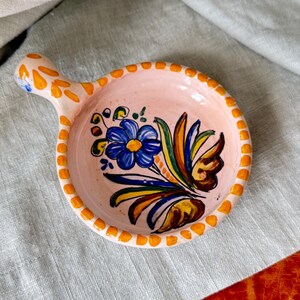 May include: A small, round, pink ceramic dish with a handle. The dish is decorated with a blue flower and colorful leaves. The rim is lined with orange dots. The handle is decorated with orange leaf shapes.