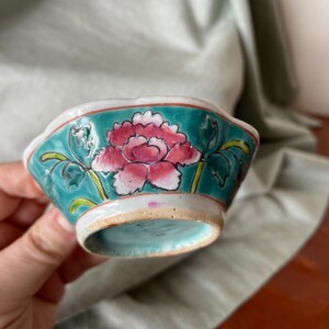 Puede incluir: Un peque&ntilde;o cuenco de cer&aacute;mica blanca con un borde azul verdoso y un dise&ntilde;o floral. El cuenco presenta una flor rosa y blanca con hojas verdes.