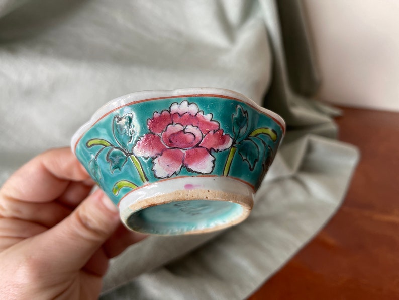 Puede incluir: Un peque&ntilde;o cuenco de cer&aacute;mica blanca con un borde azul verdoso y un dise&ntilde;o floral. El cuenco presenta una flor rosa y blanca con hojas verdes.