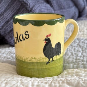 May include: A ceramic mug with a yellow background, featuring a black rooster illustration and the name "olas" in black script. The mug has a green scalloped rim and a green band at the base. The handle is yellow.