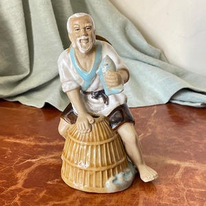 May include: A ceramic figurine of an elderly man holding a fish and leaning on a woven basket. The man wears a white shirt with blue trim, brown shorts, and a black belt. The basket is light brown with a blue base.