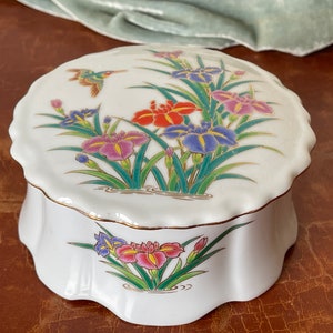 May include: White ceramic trinket box with a scalloped edge, adorned with a floral design featuring irises in shades of red, purple, and blue, with a hummingbird. The box has gold trim.