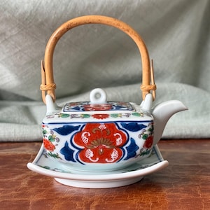 May include: A small, square ceramic teapot with a bamboo handle and matching saucer. The teapot features a floral design in red, blue, and green. The handle is made of woven bamboo. The teapot sits on a white saucer.