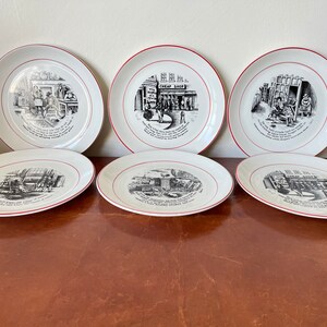 May include: Six white ceramic plates with red trim and black and white illustrations of scenes from a city street. The illustrations include a "Cheap Shop" sign, a man with a cart, and a man with a ladder.