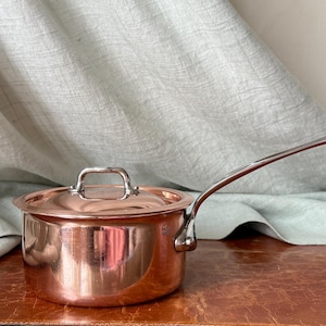 May include: A polished copper saucepan with a matching lid and a long, slender stainless steel handle. The side of the pan features a small, stamped mark that reads Made in France. The metal surface is reflective and smooth throughout.