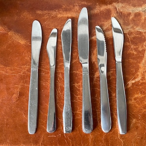 May include: Seven silver butter knives laid out on a brown leather surface.