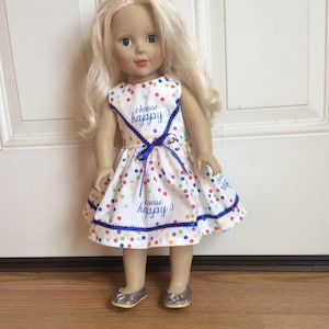 Puede incluir: Un vestido blanco con lunares de colores y ribete azul. El vestido tiene la frase "choose happy" bordada en el corpiño y el dobladillo. Una muñeca lleva el vestido y tiene el pelo rubio y los ojos azules.