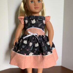 May include: A doll wearing a black and pink dress with a cat print. The dress has white ruffles on the shoulders and a white ribbon at the waist. The doll has blonde hair and blue eyes.