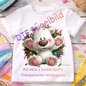 Könnte beinhalten: Ein weißes T-Shirt mit kurzen Ärmeln und einem Motiv eines weißen Bären, der in einem Weihnachtskranz mit bunten Lichtern sitzt. Der Text DTF Bügelbild, Für Helle & Dunkle Stoffe und Transparenter Hintergrund ist auf dem T-Shirt aufgedruckt.
