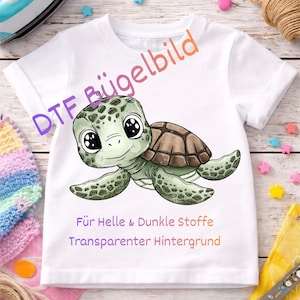 Könnte beinhalten: Weißes T-Shirt mit einem Cartoon-Schildkröten-Design. Die Schildkröte ist grün und braun mit großen Augen. Auf dem Shirt steht der Text: "Für Helle & Dunkle Stoffe Transparenter Hintergrund" und "DTF Bügelbild".