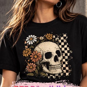 Könnte beinhalten: Ein schwarzes T-Shirt mit einem Grafikdruck eines menschlichen Schädels, umgeben von Gänseblümchen und einem Karomuster. Unten im Bild befindet sich ein rosa Banner mit dem Text DTF Bügelbilder Dunkle Hintergründe.