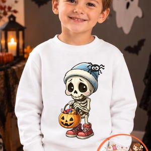 Könnte beinhalten: Ein weißes Langarm-Sweatshirt mit einer grafischen Illustration eines Cartoon-Skeletts, das eine blaue Mütze und rote Turnschuhe trägt und einen mit bunten Süßigkeiten gefüllten Kürbis-Eimer hält, mit einer kleinen Spinne auf der Mütze.