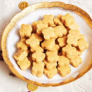 May include: A white plate with a gold rim, filled with small, star-shaped, light brown cookies. The plate is on a white tablecloth with gold coins scattered around it.