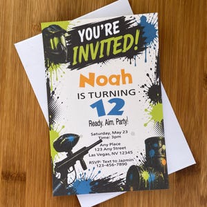 May include: A paintball-themed invitation with the text "YOU'RE INVITED!" and "Noah IS TURNING 12". The invitation includes a paintball gun graphic, paint splatters, and RSVP information. The invitation is on a wooden surface.