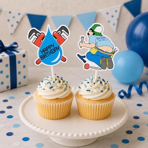 Peut inclure: Deux cupcakes avec glaçage blanc et vermicelles bleus sur un support blanc. Une décoration est une goutte d'eau bleue avec des clés à pipe rouges et le texte HAPPY BIRTHDAY. L'autre est un plombier en chemise bleue et casquette avec outils.
