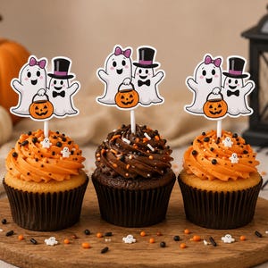 May include: Three cupcakes topped with orange and chocolate frosting, decorated with sprinkles and white ghost-shaped candies. Each cupcake features a paper topper showing two cartoon ghosts, one with a pink bow and one with a black top hat.