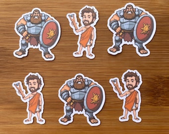 May include: A collection of six stickers featuring cartoon illustrations. Three stickers depict a muscular figure in armor with a shield, while the other three show a person in an orange toga holding a slingshot. The stickers are on a wooden surface.