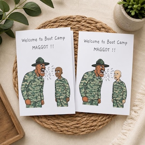 Könnte beinhalten: Zwei identische weiße Grußkarten mit einer Cartoon-Illustration eines Drill-Sergeants in Tarnuniform, der einen kahlköpfigen Rekruten in Tarnuniform anschreit. Der Text lautet Welcome to Boot Camp MAGGOT !! in schwarzer Schrift.