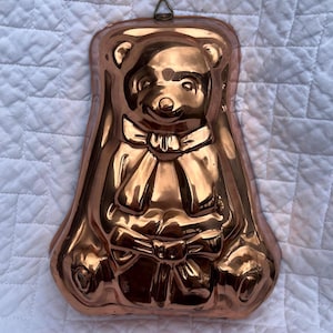 May include: A copper-colored bear-shaped mold with a bow tie and a bow on the bottom. The mold has a hanging loop at the top. The bear's features are detailed, with visible eyes, nose, and ears. The mold is likely for baking or making decorative items.