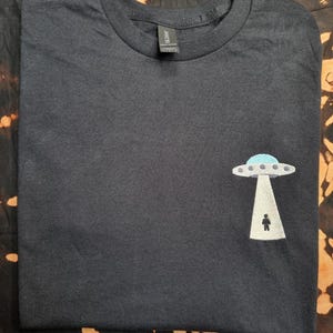 Könnte beinhalten: Ein schwarzes T-Shirt mit Rundhalsausschnitt und einer Stickerei auf der unteren rechten Seite. Das Motiv zeigt ein graues und hellblaues UFO, das über einem weißen Lichtstrahl schwebt, mit einer kleinen schwarzen Figur im Lichtkegel.