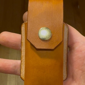 May include: A handcrafted, light brown leather case with a flap closure secured by a brass button. The case has a rectangular shape with rounded edges and a contrasting light-colored stitching.